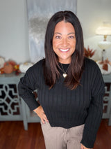 Black ribbed long-sleeve sweater worn with beige pants, featuring a crew neckline and fitted silhouette. Model is wearing a gold pendant necklace and smiling at the camera in a bright, neutral interior setting.