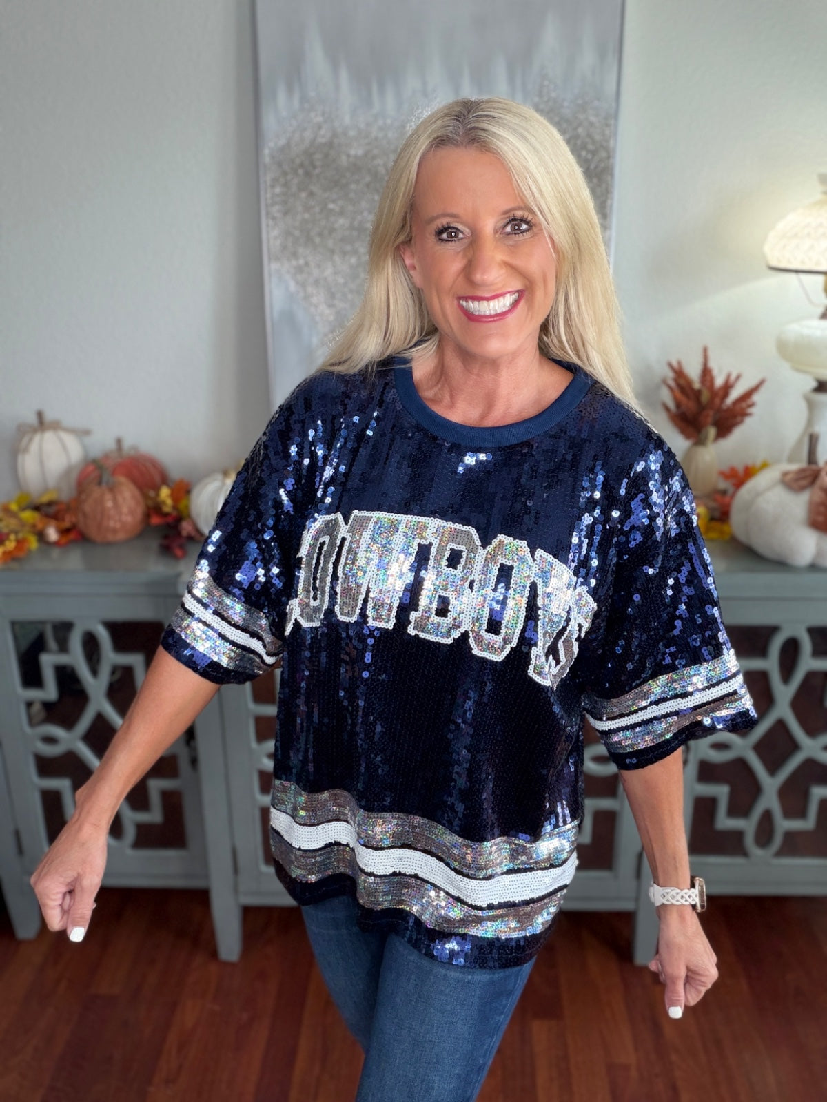 Woman wearing a navy blue sequined Cowboys football jersey top with white lettering and stripe detailing on the sleeves, paired with blue jeans, in a home interior setting.