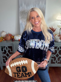 A woman wears a navy blue sequined short-sleeve top with "Cowboys" text across the chest, paired with blue jeans. She holds a brown football-shaped sign that reads "GO TEAM!"