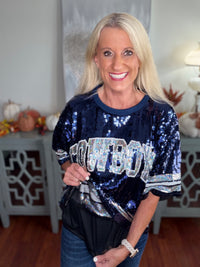 A woman wearing a navy blue sequined short-sleeve top with white and silver "COWBOYS" text across the chest, paired with blue jeans. The sequined garment features sparkly embellishments and a casual, sporty aesthetic.