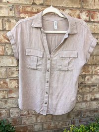 A light mauve linen short-sleeve button-up shirt displayed on a white hanger against a brick wall. The shirt features two chest pockets and a collared neckline with rolled sleeves.