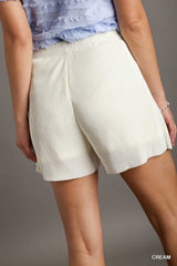 A woman wearing cream-colored ribbed shorts with a relaxed fit, paired with a lavender textured top. The shorts feature a vertical ribbed texture and reach mid-thigh length.