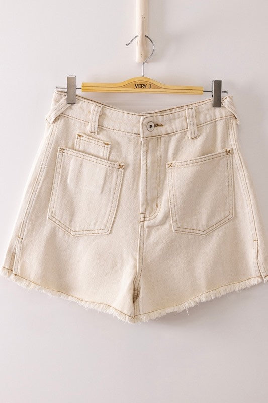Cream-colored denim shorts with high waist and raw frayed hem, featuring visible stitching details and multiple pockets, displayed on a wooden hanger against a neutral background.