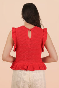 A woman wearing a bright red sleeveless knit top with ruffled flutter sleeves and a keyhole cutout on the back, paired with a cream-colored lace skirt.