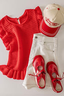 A red textured knit sweater with ruffled short sleeves and hem is styled with cream-colored pants, red and white canvas sneakers, and a red and cream baseball cap featuring embroidered text.