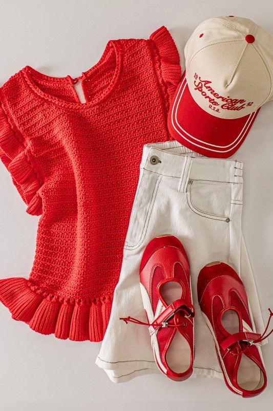 A red textured knit sweater with ruffled short sleeves and hem is styled with cream-colored pants, red and white canvas sneakers, and a red and cream baseball cap featuring embroidered text.