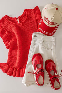 A red textured knit sweater with ruffled short sleeves and hem is styled with cream-colored pants, red and white canvas sneakers, and a red and cream baseball cap featuring embroidered text.