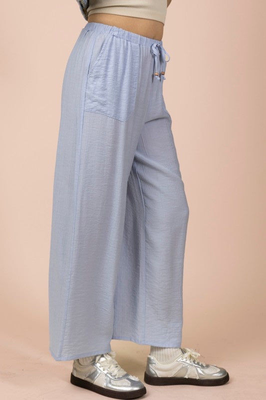A model wearing light blue wide-leg pants with a drawstring waist and side pocket, paired with a cream-colored ribbed crop top and white platform sneakers against a beige background.