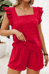 A woman wearing a bright red textured romper with ruffled flutter sleeves and a square neckline, paired with layered necklaces and hoop earrings.