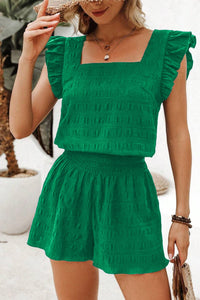 A woman wearing a vibrant green two-piece set consisting of a square-neckline crop top with ruffled cap sleeves and matching high-waisted shorts with an elastic waistband, accessorized with a straw hat and beaded bracelets.