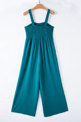 A teal sleeveless jumpsuit with a smocked bodice and wide-leg pants, displayed on a wooden hanger against a white background.