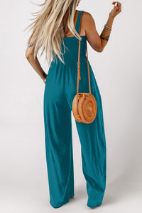 A woman wearing a teal sleeveless jumpsuit with wide-leg pants and a halter neckline, accessorized with a woven rattan crossbody bag with an orange strap and layered bracelets.