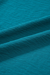 Close-up of a teal blue textured fabric with subtle horizontal ribbing and diagonal folds, showing the soft, matte finish and drape of the material.