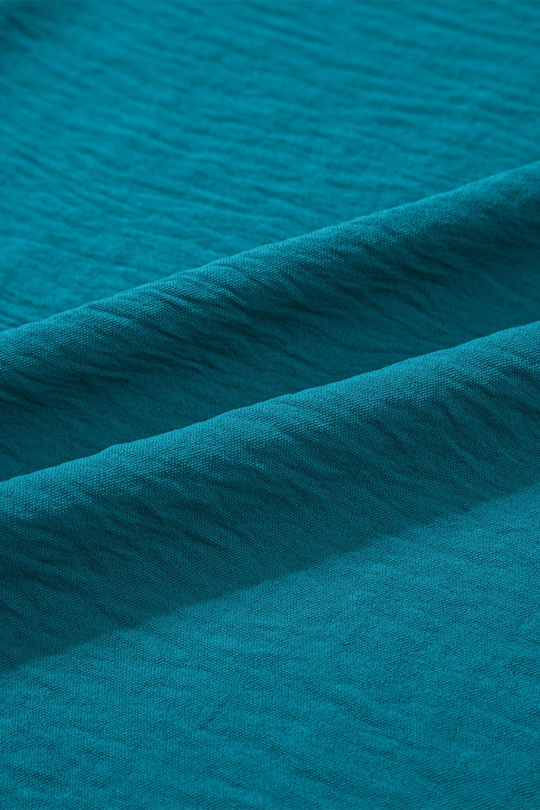 Close-up of a teal blue textured fabric with subtle horizontal ribbing and diagonal folds, showing the soft, matte finish and drape of the material.