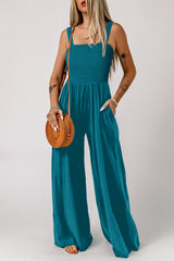 A woman wearing a teal sleeveless jumpsuit with a smocked bodice and wide-leg pants, paired with a woven tan crossbody bag and beaded bracelets.