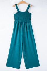A teal sleeveless jumpsuit with a smocked bodice and wide-leg pants, displayed on a wooden hanger against a white background.