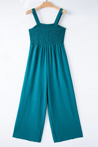 A teal sleeveless jumpsuit with a smocked bodice and wide-leg pants, displayed on a wooden hanger against a white background.