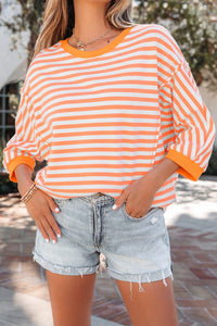 A woman wears an orange and white striped long-sleeve top with rolled cuffs, paired with light blue distressed denim shorts. She accessorizes with a silver chain necklace and beaded bracelets.