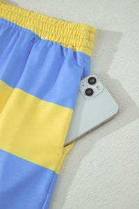 A white smartphone with dual rear cameras sits next to blue and yellow striped athletic shorts with a yellow elastic waistband, displayed on a light gray textured surface.