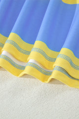 Close-up of a striped beach towel featuring blue and yellow horizontal stripes with gray decorative trim details, laid on sand showing its textured fabric weave.