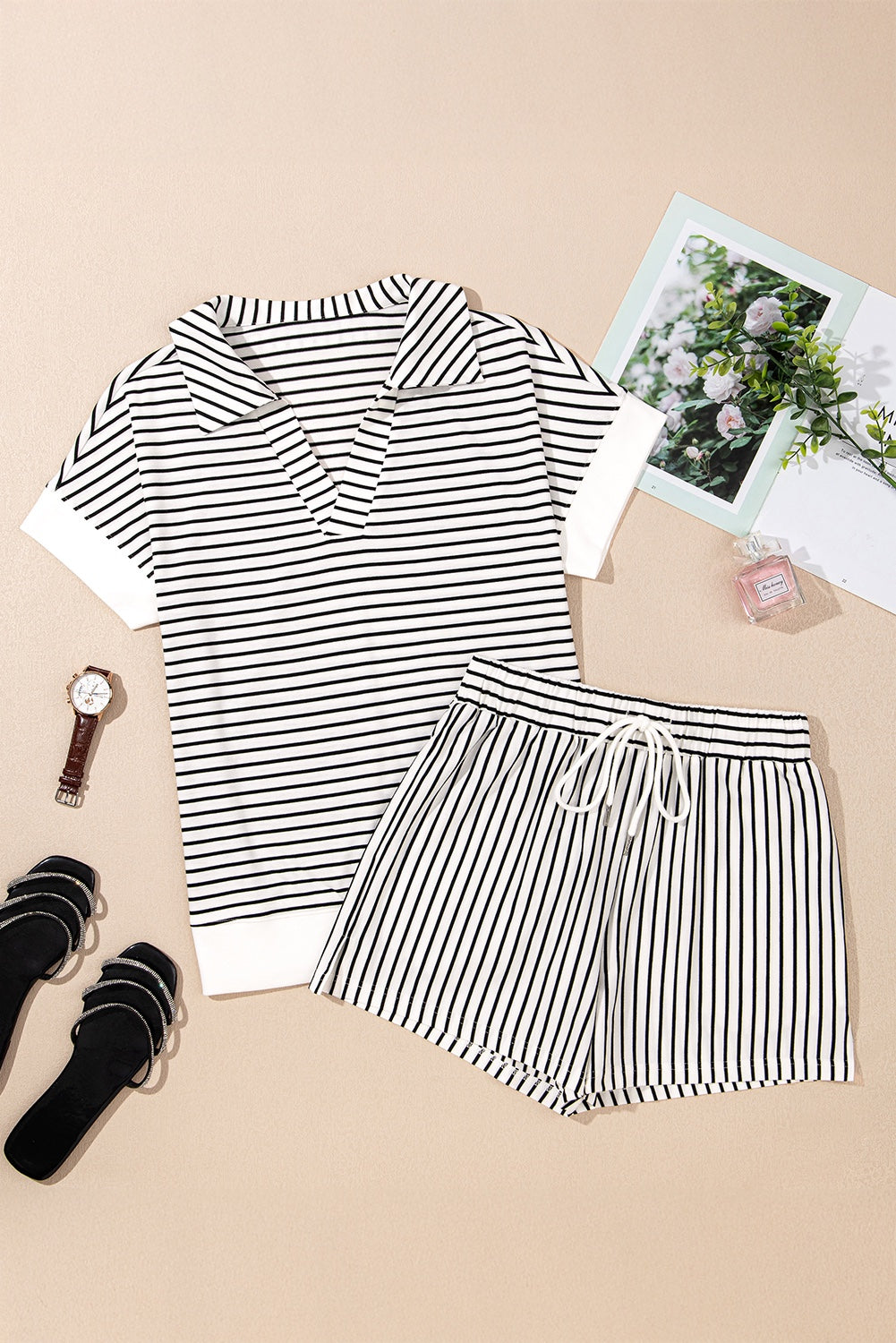 A black and white striped short-sleeve t-shirt and matching striped shorts set laid flat on a beige background, styled with black sandals, a watch, and decorative accessories including photos and a perfume bottle.
