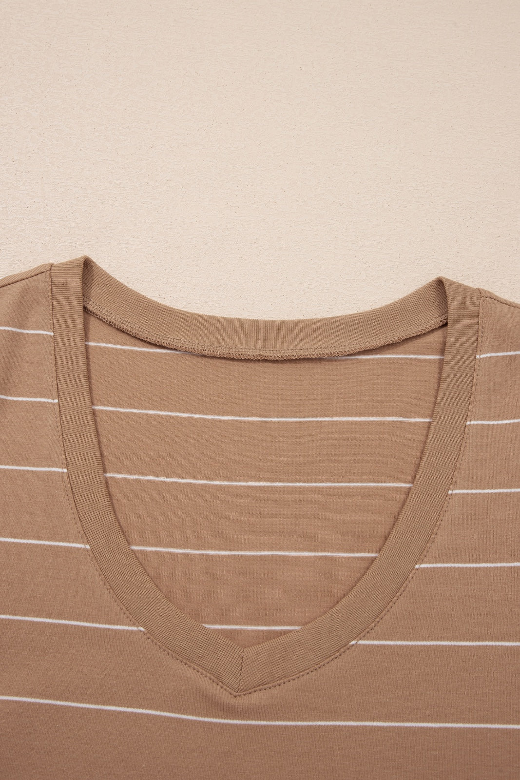 A tan ribbed tank top with white horizontal stripes, featuring a V-neckline and fitted shoulders, displayed flat against a neutral beige background.