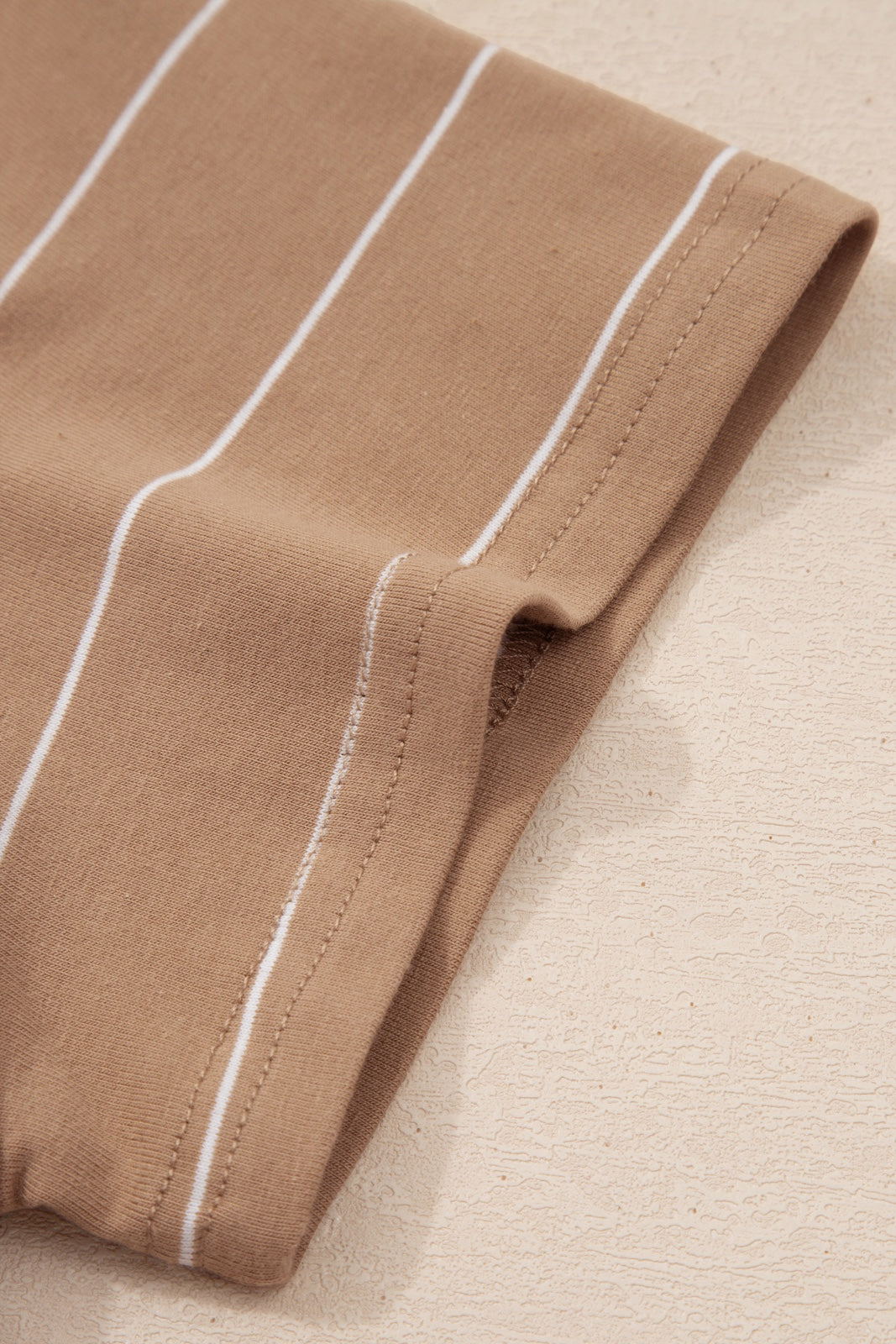Close-up of a beige striped knit fabric with white vertical stripes and a rolled edge detail, showing the garment's texture and construction seaming.
