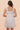 A woman wearing a light lavender sleeveless dress with a square neckline, smocked bodice, and delicate eyelet texture throughout. The dress features ruffled shoulder straps and falls to knee length with a gathered waist.