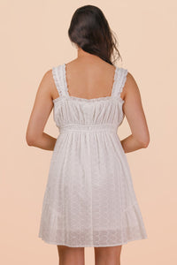 A woman wearing a light lavender sleeveless dress with a square neckline, smocked bodice, and delicate eyelet texture throughout. The dress features ruffled shoulder straps and falls to knee length with a gathered waist.