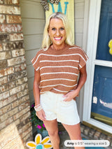 Woman wearing a rust-orange and white striped short-sleeve knit top with white distressed denim shorts, standing on a porch with brick accents.