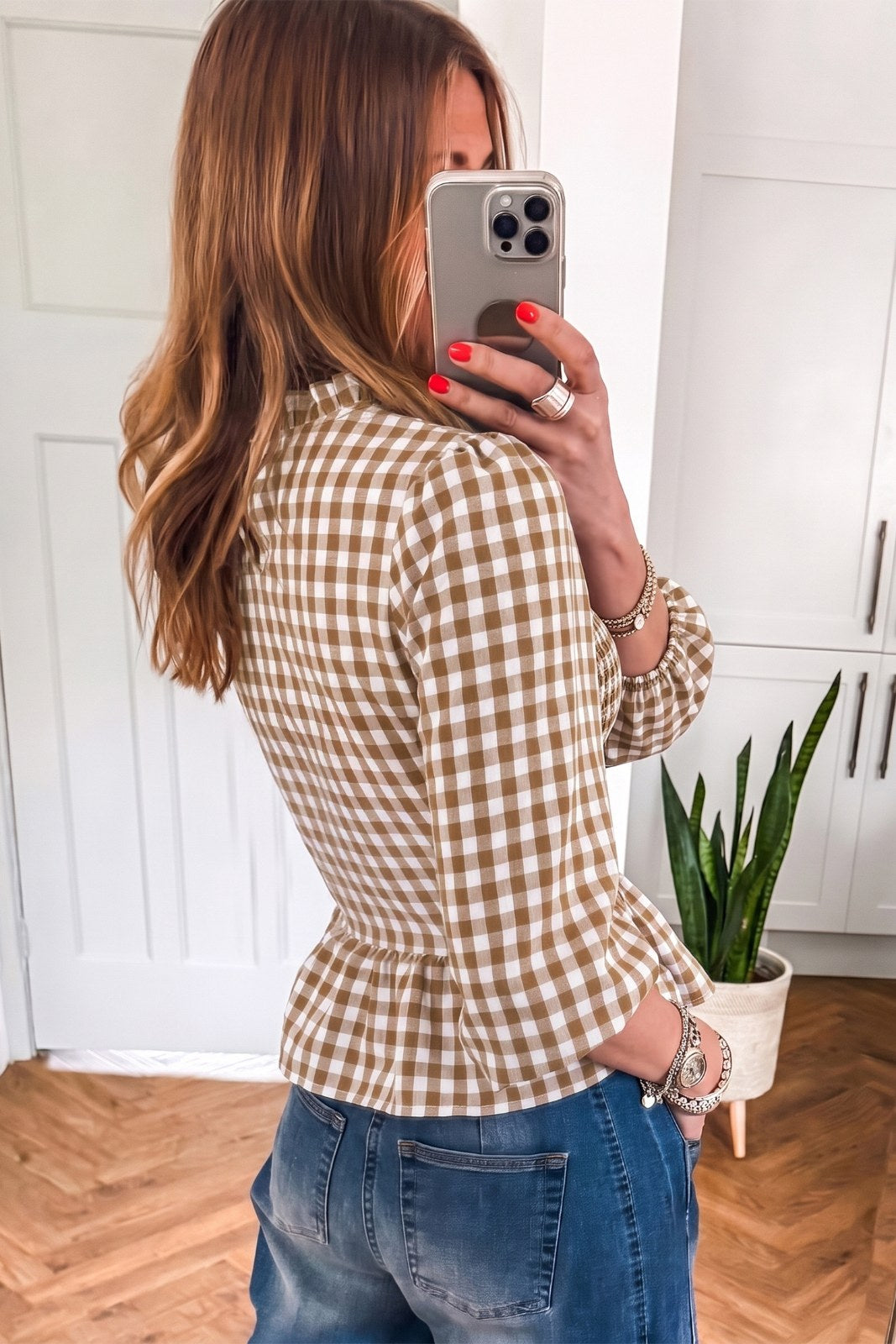 Woman wearing a tan and white gingham check button-up shirt paired with blue jeans, taking a mirror selfie. The shirt features long sleeves and a fitted silhouette.