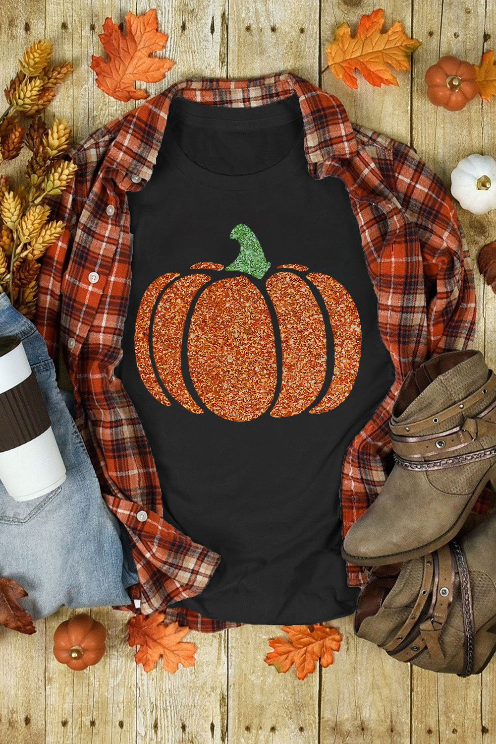 Black sleeveless tank top featuring a glittery orange pumpkin with a green stem, displayed flat lay with a plaid flannel shirt, autumn leaves, small pumpkins, boots, and fall decorations on a wooden background.
