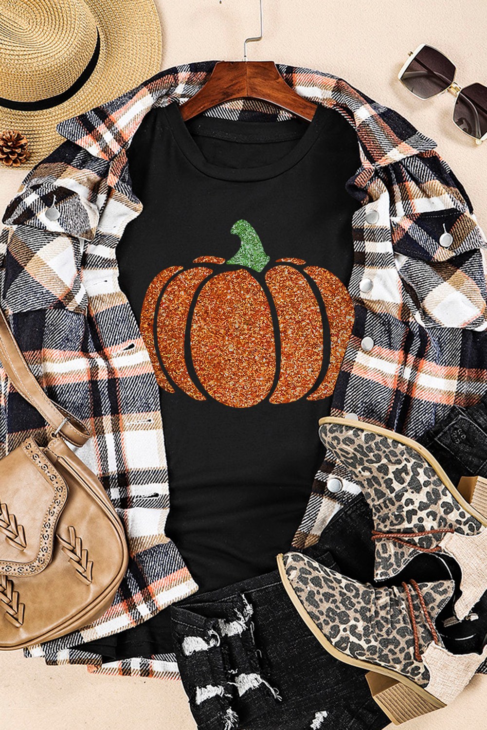 A black short-sleeve t-shirt featuring a glittery orange pumpkin with a green stem, displayed flat lay with a plaid shirt, straw hat, leopard print boots, tan bag, and sunglasses.