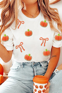 A white short-sleeved t-shirt featuring a repeating pattern of orange pumpkins and red bows, styled with light blue jeans and a matching orange pumpkin-print cup.