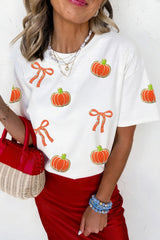 A woman wearing a white short-sleeve t-shirt featuring an all-over print of orange pumpkins and red bows, styled with red pants and layered silver necklaces.