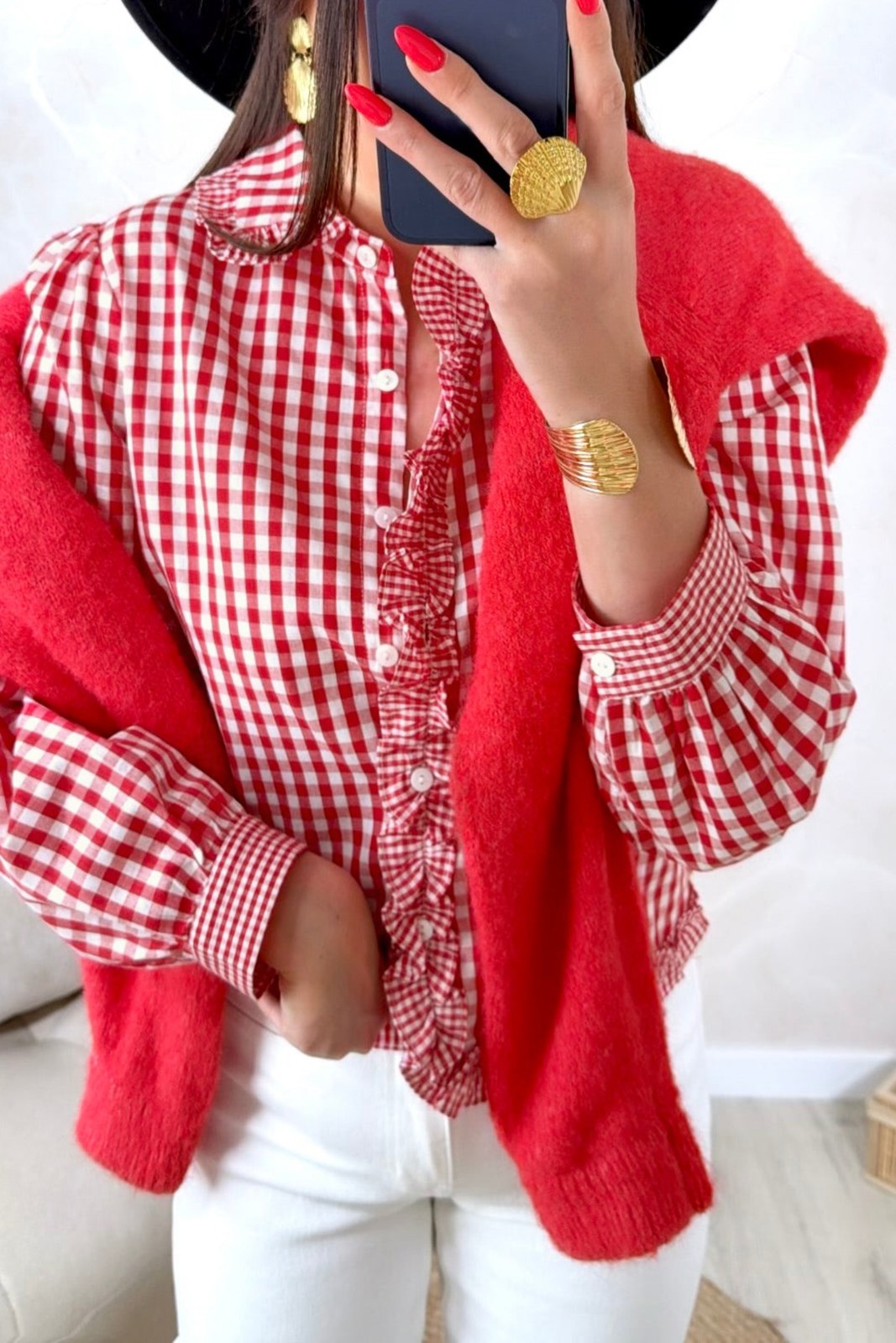 A red and white gingham check blouse with ruffled front placket and voluminous sleeves, paired with a solid red cardigan. The outfit is styled with gold circular earrings and red nail polish.