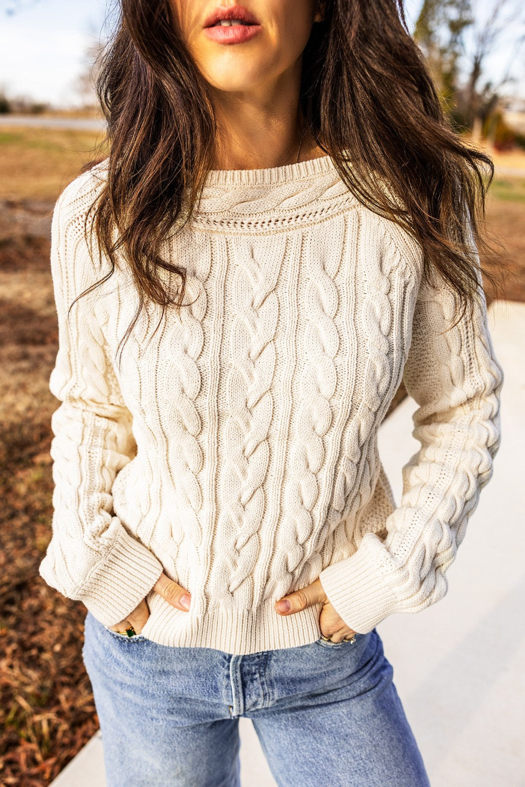 A woman wearing a cream cable-knit sweater with a crew neckline and long sleeves, paired with light blue denim jeans, standing outdoors in a rural setting.