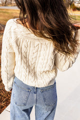 A person wearing a cream-colored cable knit cropped cardigan over blue denim jeans, shown from behind with long dark hair flowing over one shoulder.