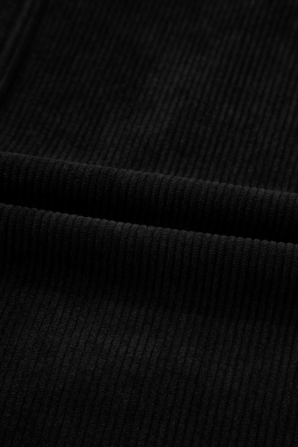 Close-up of black ribbed knit fabric showing vertical texture and subtle folds in the material. The garment features a classic ribbed knit pattern in solid black.