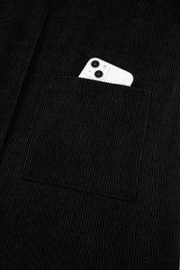 A white smartphone with dual cameras peeks out from the breast pocket of a black ribbed knit sweater or cardigan, showcasing the garment's functional pocket design.