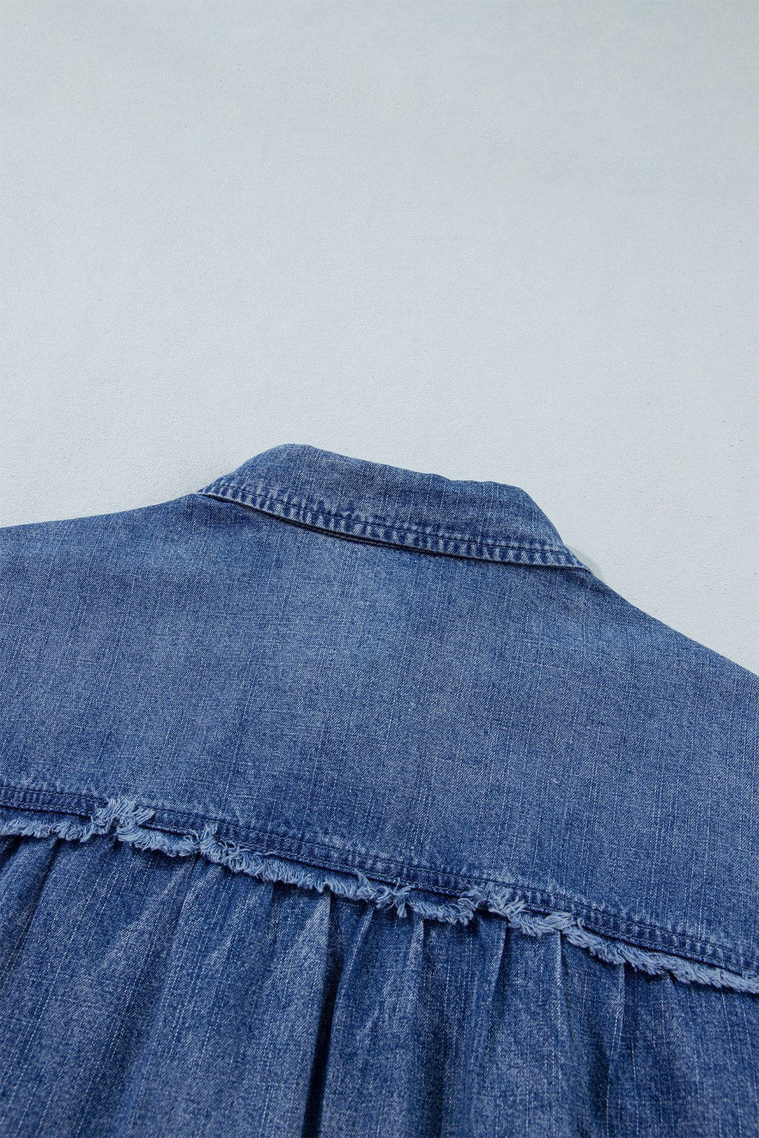 Close-up of a blue denim garment showing the back yoke and upper back panel with visible stitching and a ruched or gathered detail along the lower edge.