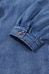 Close-up of a blue denim shirt cuff featuring a gathered fabric design and a copper-toned button, showing the classic texture and weave of chambray or light denim material.