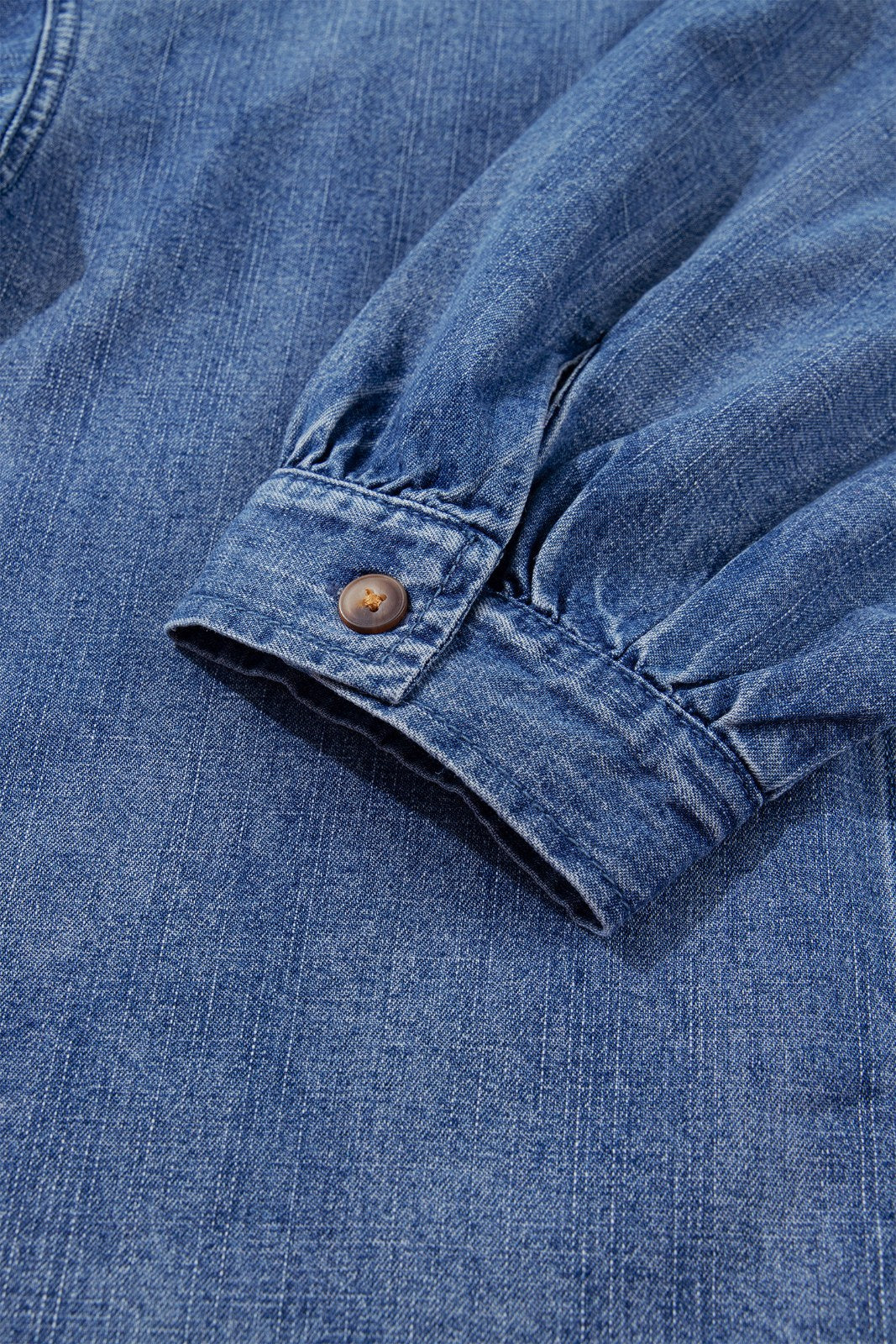 Close-up of a blue denim shirt cuff featuring a gathered fabric design and a copper-toned button, showing the classic texture and weave of chambray or light denim material.