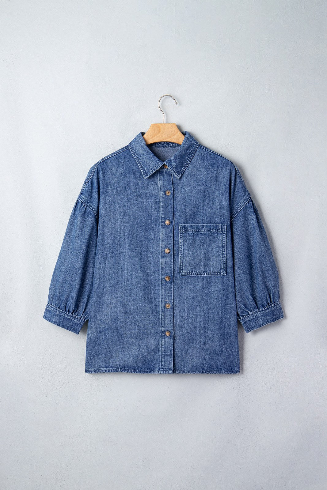 A blue denim button-up shirt displayed on a wooden hanger against a white background, featuring a classic collar, chest pocket, and three-quarter length sleeves with rolled cuffs.