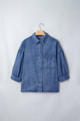 A blue denim button-up shirt displayed on a wooden hanger against a white background, featuring a classic collar, chest pocket, and three-quarter length sleeves with rolled cuffs.