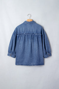 Back view of a blue denim shirt on a wooden hanger, featuring gathered fabric at the waist, long sleeves with cuffed hems, and a button-front closure against a light gray background.