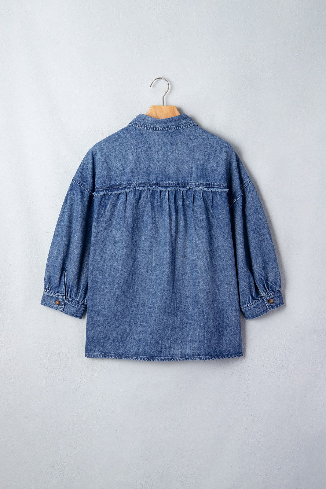 Back view of a blue denim shirt on a wooden hanger, featuring gathered fabric at the waist, long sleeves with cuffed hems, and a button-front closure against a light gray background.