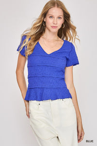 A woman wearing a bright blue short-sleeve v-neck top with horizontal textured stripes and a ruffled peplum hem, paired with cream-colored pants.