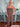 A woman wears a brown strapless jumpsuit with vertical pleating and wide-leg pants. The outfit features a gathered neckline and is styled with layered silver jewelry and floral sandals.