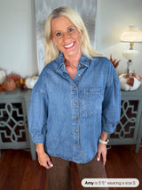 A woman wearing an oversized denim button-up shirt in medium blue with a chest pocket, styled with brown pants. The shirt features long sleeves and a classic collared design.